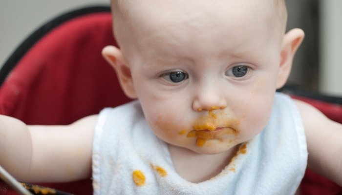 A 6-month-old baby in a high chair with some food on his face and bib.