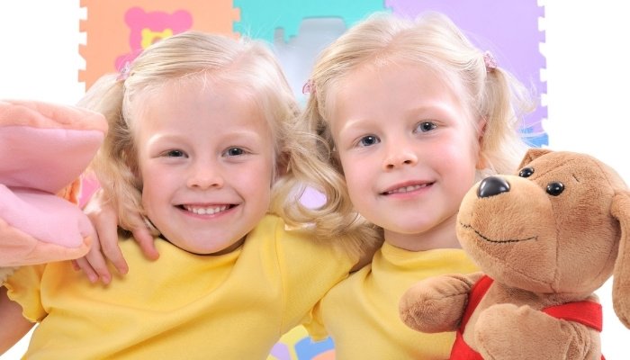 Blonde twin sisters in yellow shirts holding stuffed animals.