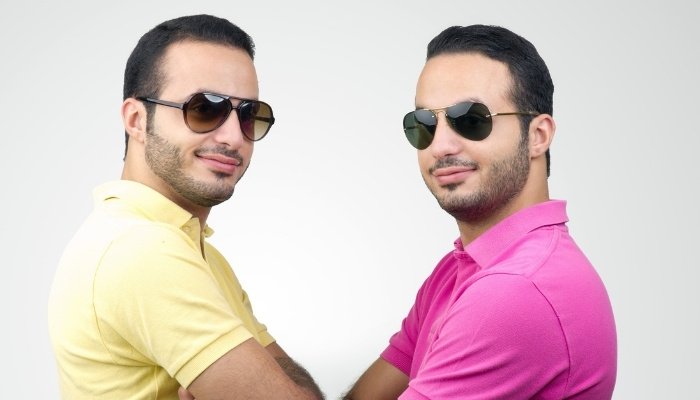 Identical twin men wearing sunglasses and pink and yellow shirts facing each other with arms crossed.