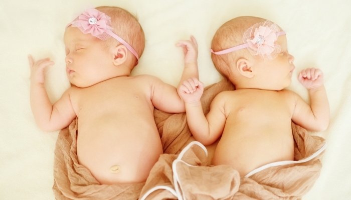 Newborn twin girls sleeping together with light-brown blanket.