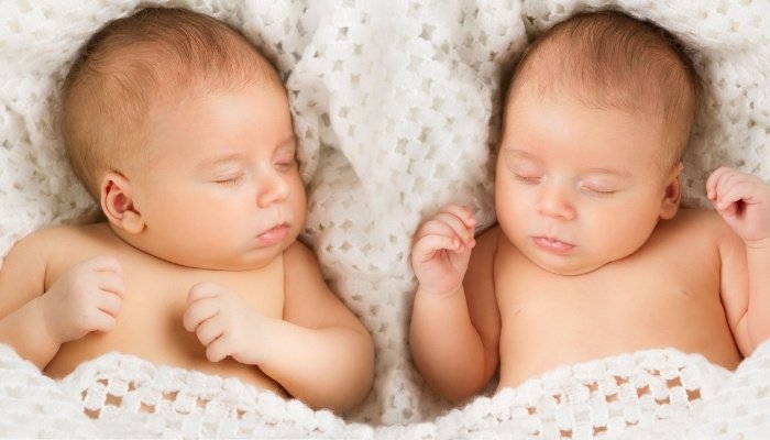 Newborn twins sleeping side by side under a white crochet blanket.