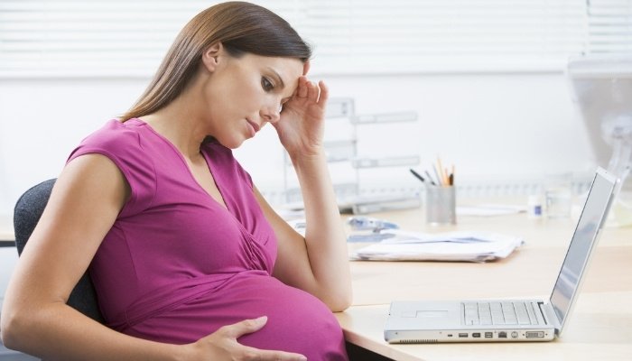 A pregnant woman sitting at work in front of a laptop looking worried.