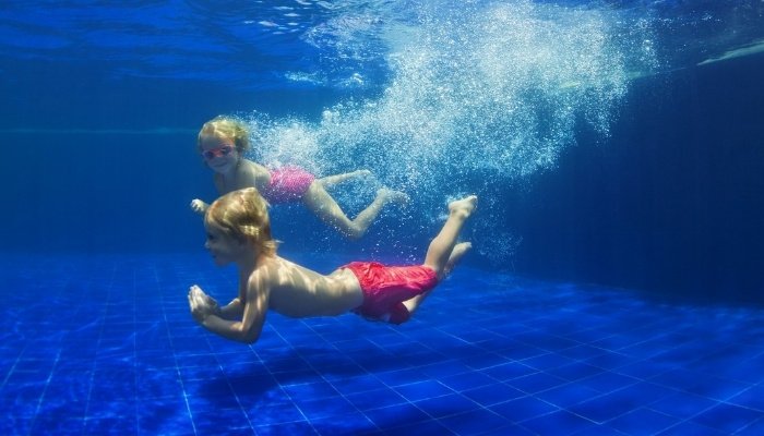 Twin brothers in red swimsuits swimming underwater in a pool.