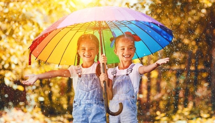 Two twin sisters enjoying a rain shower while under a large rainbow umbrella.