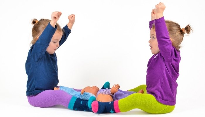 Twin sisters sitting on the floor with arms raised and two dolls lying between them.