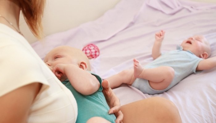 A twin mom breastfeeding one baby while the other lies on the bed in front of her.