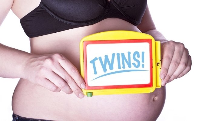 A pregnant woman holding a sign that says Twins in front of her belly.