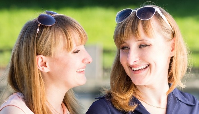 Adult twin sisters with sunglasses perched on top of their heads talking and smiling while outside.