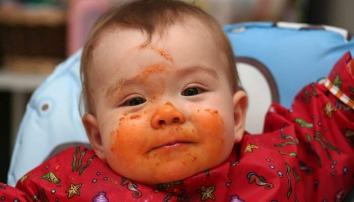 A cute baby wearing a red smock with spaghetti sauce smeared on her face.