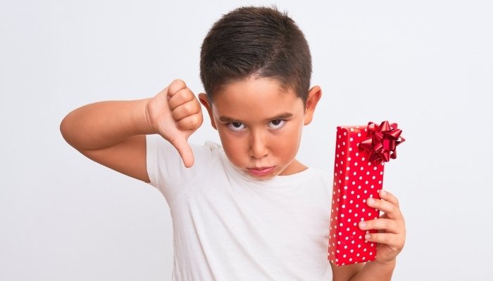 A young boy looking ungrateful and giving the thumbs down sign while holding a wrapped present.