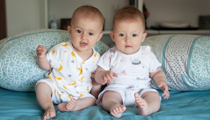 Fraternal twin girl babies sitting on bed in front of two pillows.