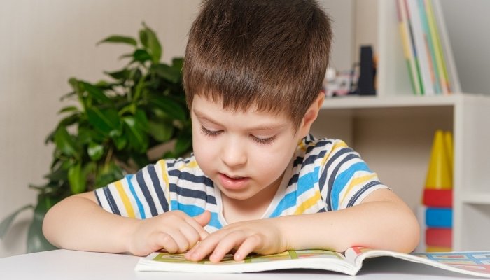 A 4-year-old in a colorful striped shirt sitting at a table reading a book.