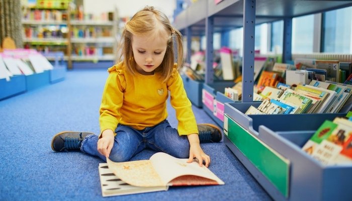 A little girl in a yellow shirt sitting on the floor at a library looking at a book.