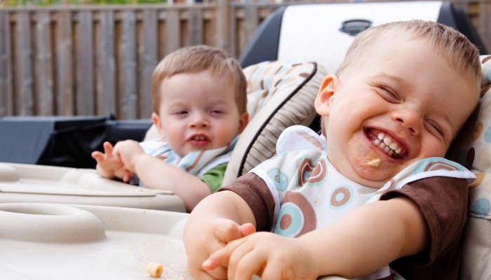 Twin brothers laughing and eating in their highchairs.
