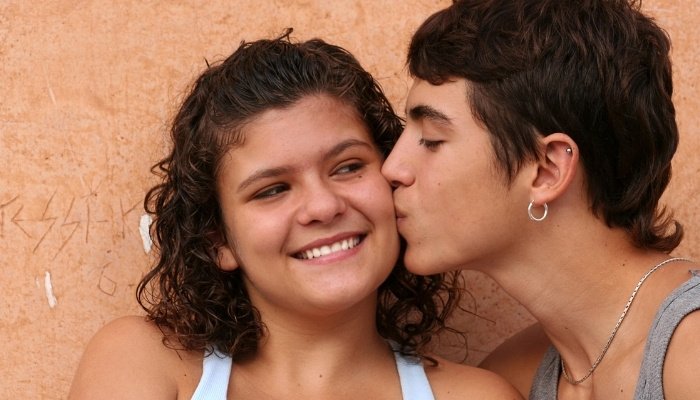 A teen boy kissing a young teen girl on the cheek.