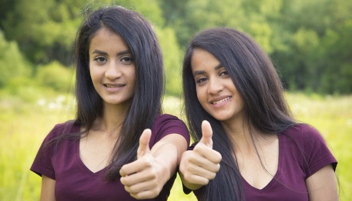 Adult twin sisters in purple shirts outdoors giving the thumbs up sign.