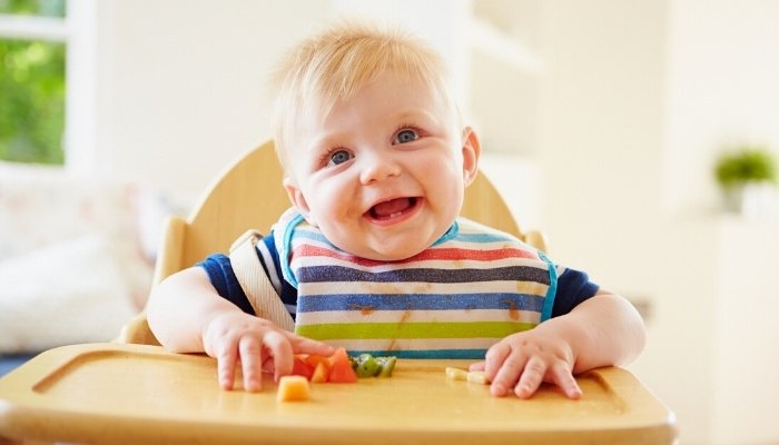 A baby boy in a striped shirt feeding himself cut fruit in a high chair.