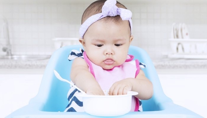 A cute baby girl wearing a bow on her head sitting in a high chair and reaching into a bowl for food.