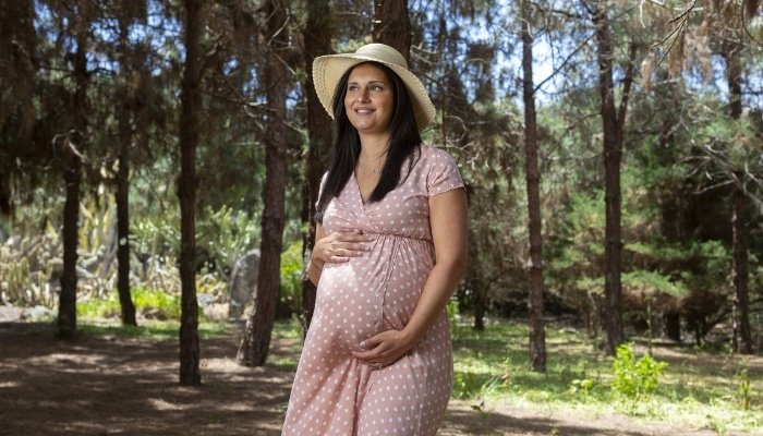 A smiling, happy pregnant woman in a dress and floppy hat walking in the woods.