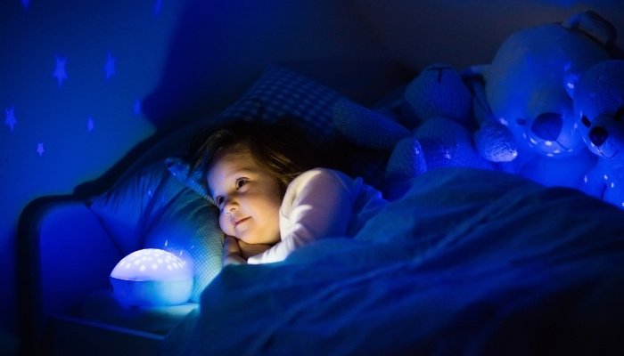 A little girl lying in her toddler bed staring at the stars on the wall projected by a nightlight.