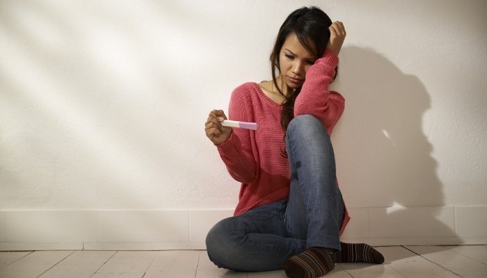 A young girl sits on the floor and stares sadly at a pregnancy test.
