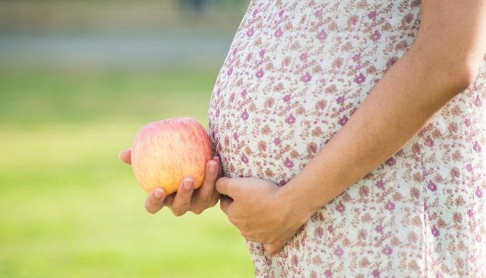 A woman cradling her pregnant belly while holding an apple.
