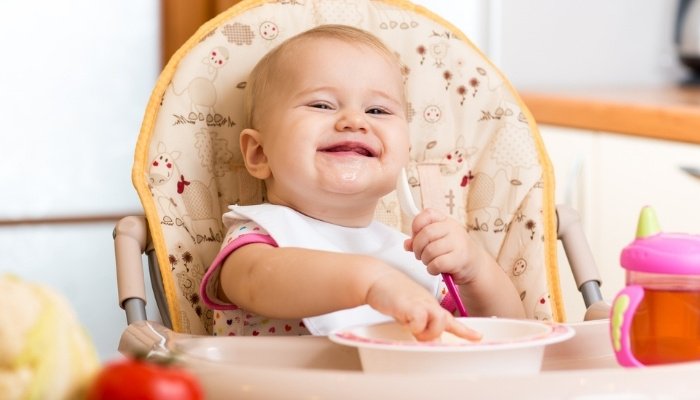 A baby girl smiling as she feeds herself from a bowl while sitting in a high chair.