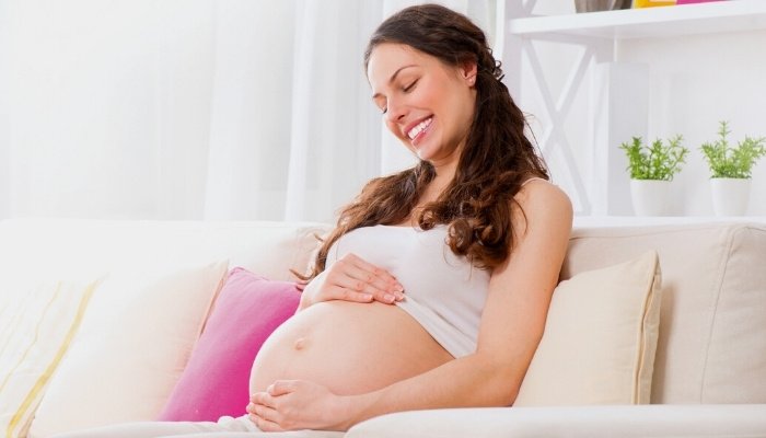 A pregnant woman sitting on her couch smiling while looking at her belly.