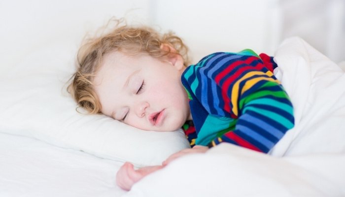 A sweet toddler with curly, wispy hair and striped pajamas asleep in bed. 