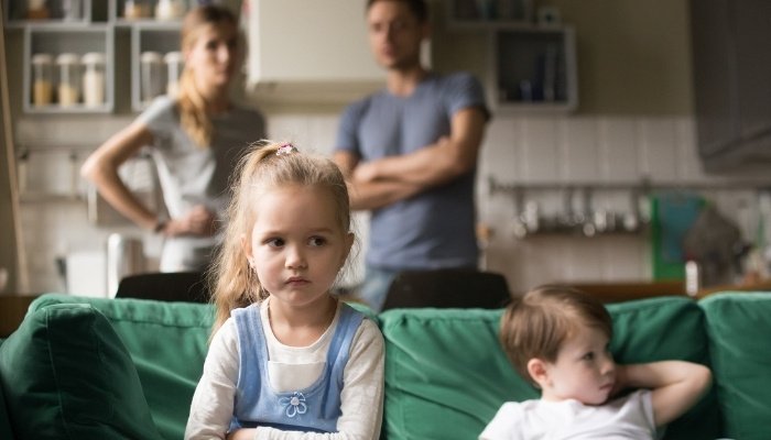 An upset girl sitting beside her brother on a couch with disappointed parents in background.