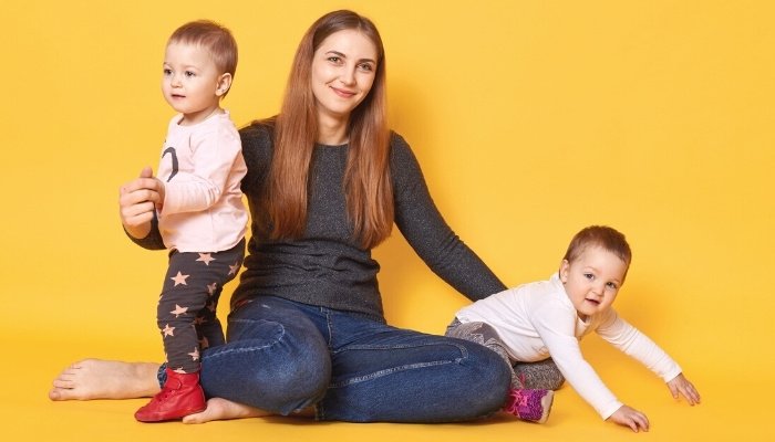 A young woman with twin toddlers against an orange background.