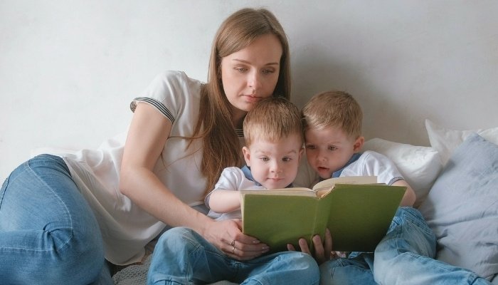 A young woman and twin toddlers lounging on bed reading a book together.
