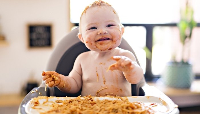 A cute baby in a high chair with spaghetti all over herself and the tray.