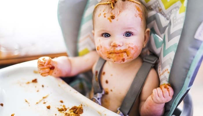 A cute baby girl sitting in a high chair eating spaghetti and making a mess.
