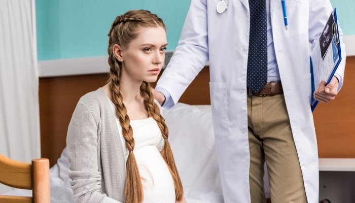 A pregnant lady sitting in a doctor's office looking devastated while the doctor tries to comfort her.