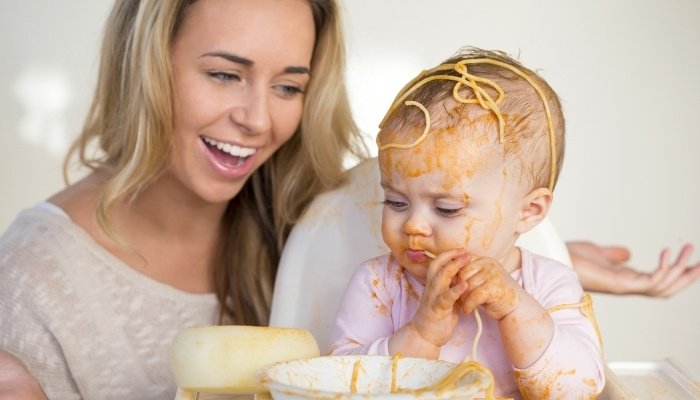 A mother watches and smiles while her baby experiments with eating spaghetti.
