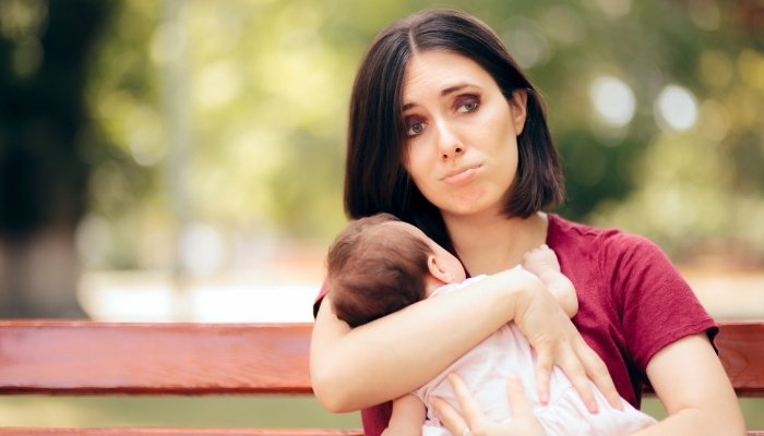 A new mom with a strange expression on her face holding her baby while sitting outside on a park bench.