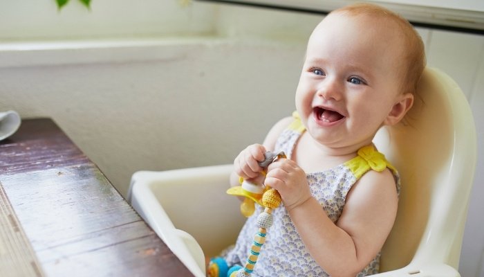 An older baby girl sitting in her high chair smiling while holding a toy.