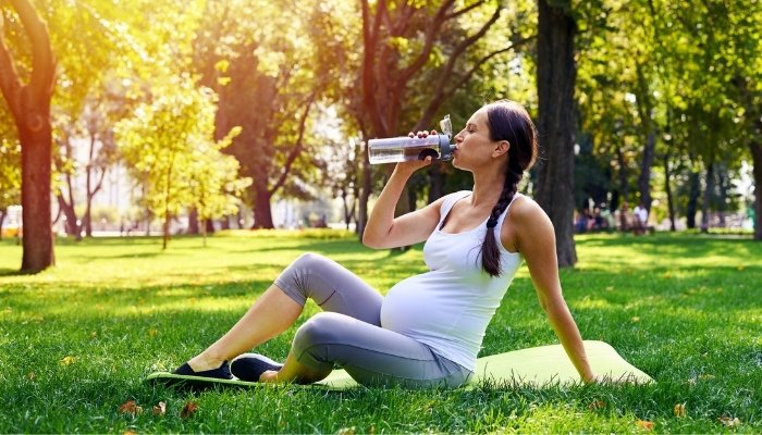 A pregnant lady dressed in exercise clothing sitting down in a park to drink water.