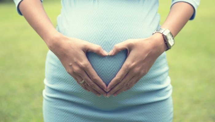 A woman in a blue dress making a heart sign over her baby bump.