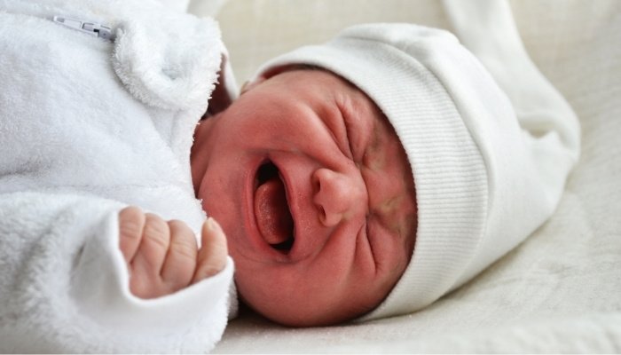 A screaming baby dressed in a white outfit and a white hat.