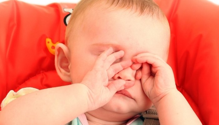 A tired baby sitting in a high chair rubbing her eyes and face.