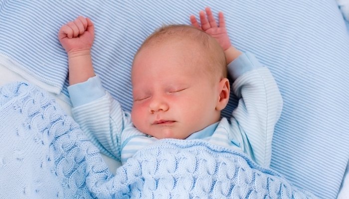 A baby boy fast asleep in his bassinet with blue sheets and a blue blanket.