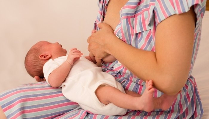 A mother holding her breast in discomfort with her baby on her lap.