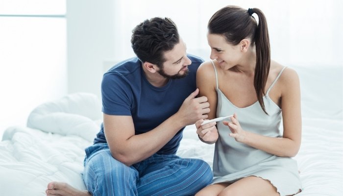 A couple sitting on their bed looking happy after taking a pregnancy test.