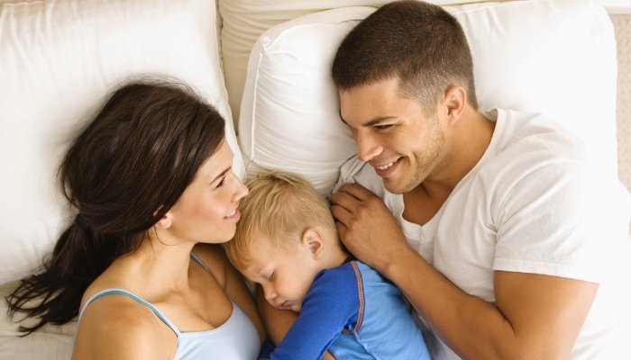 A young mom and dad lying in bed with their sleeping toddler between them.