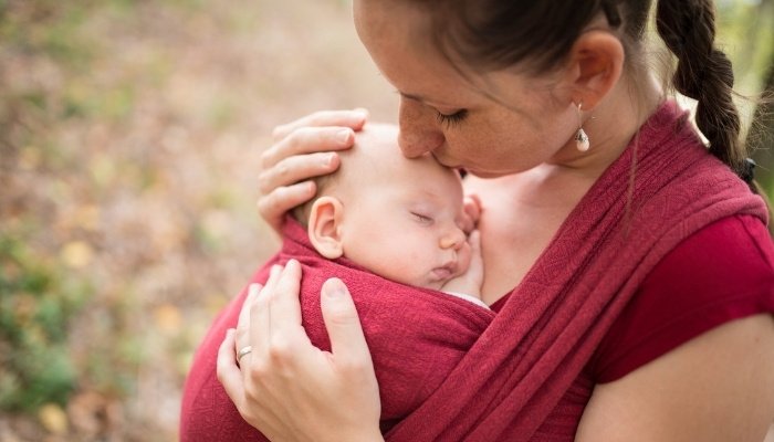 A young mother with her baby wrapped close to her chest gives her a kiss on the forehead.