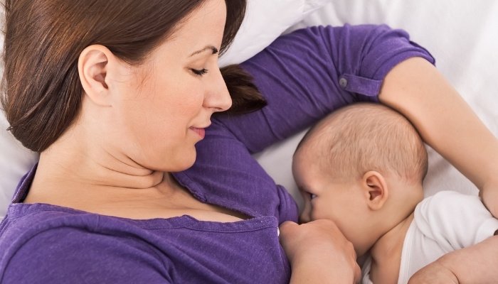 A mother in a purple shirt breastfeeding her baby while lying in bed.