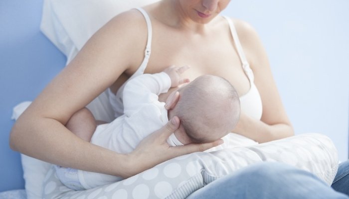 A young mother holding her baby in the football hold to nurse while sitting in bed.