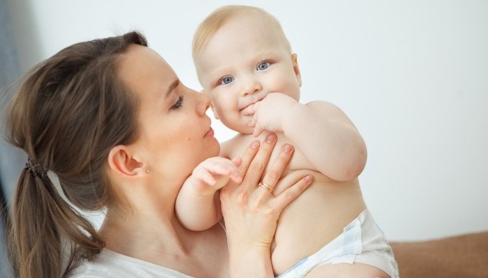 A young mother sniffing her baby's cheek while he sucks on his hand.